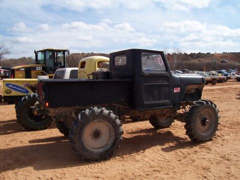 2004 Aston Martin Virage - A modified black pickup truck with large, aggressive tires is parked on a dirt lot, with other vehicles and construction equipment in the b…