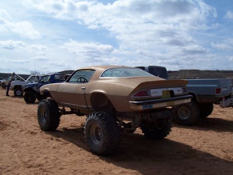 2004 Aston Martin Virage - A lifted 1970s muscle car with oversized tires sits on sandy terrain, with other trucks in the background.