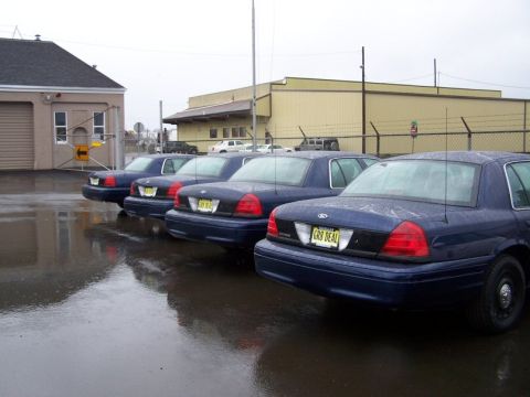 2001 Ford Crown Victoria - A line of dark blue 2001 Ford Crown Victoria police cars are parked in a wet lot under an overcast sky.