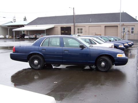 2001 Ford Crown Victoria - A row of blue 2001 Ford Crown Victoria sedans are parked outside a building on a wet day.