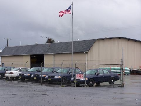 2001 Ford Crown Victoria - A row of police sedans are parked outside a building under a cloudy sky, with an American flag waving.