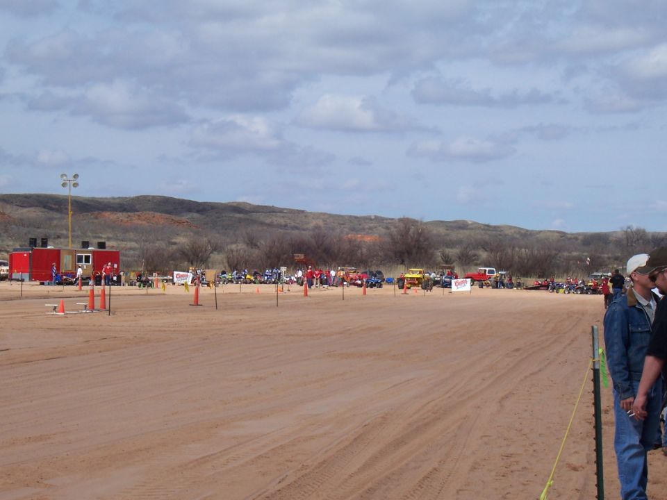 Sandy area with multiple ATVs, trucks, and people gathered under a cloudy sky
