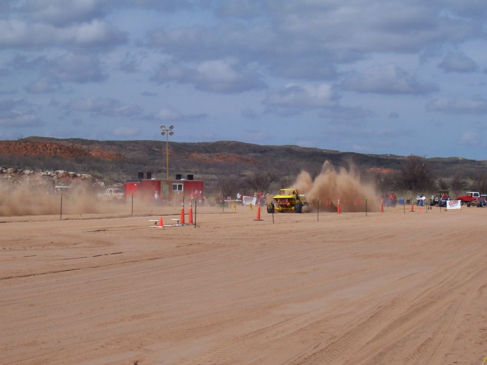 Yellow off-road buggy kicking up dust on a sandy track with spectators and buildings in the background