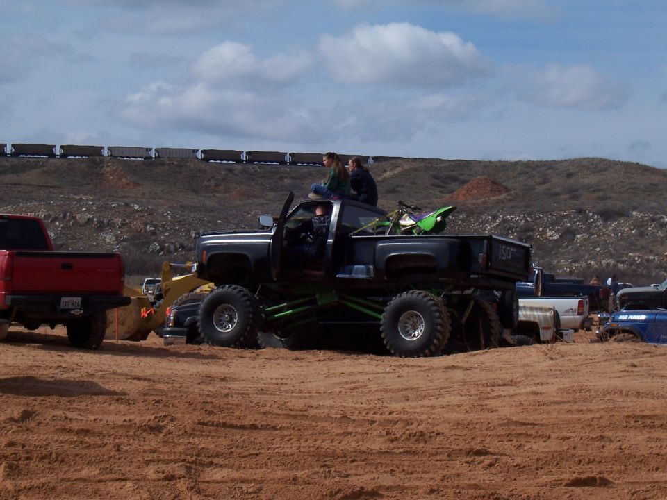 Black monster truck with green suspension, two people on top, dirt bike in bed. Other trucks in background