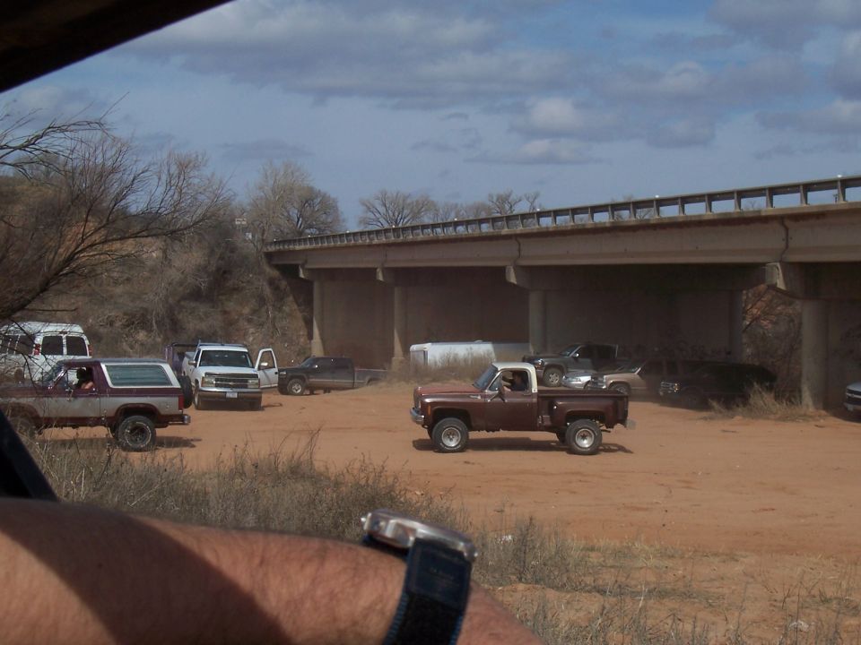 Several trucks and vans parked on a dirt lot under a concrete bridge