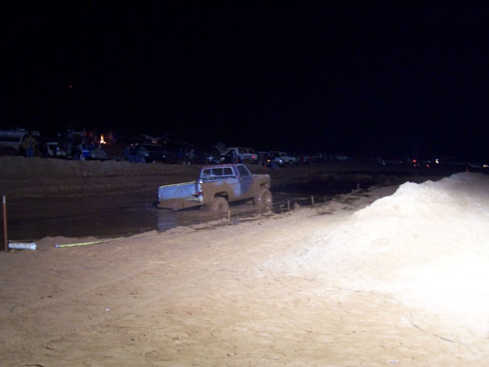 Mud-covered pickup truck stuck in a bog at night, with spectators and other vehicles in the background