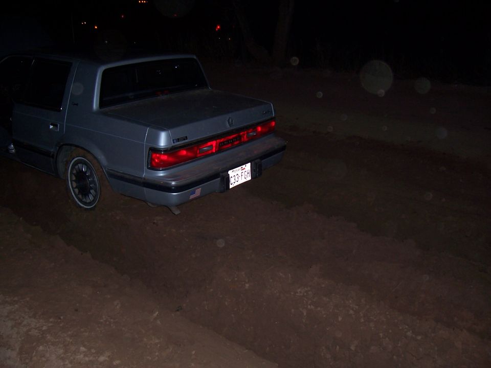 Rear of a gray sedan parked on a dirt road at night. License plate is visible