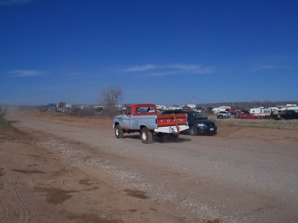 Red and gray Ford pickup truck driving on a dirt road, with a dark sedan following