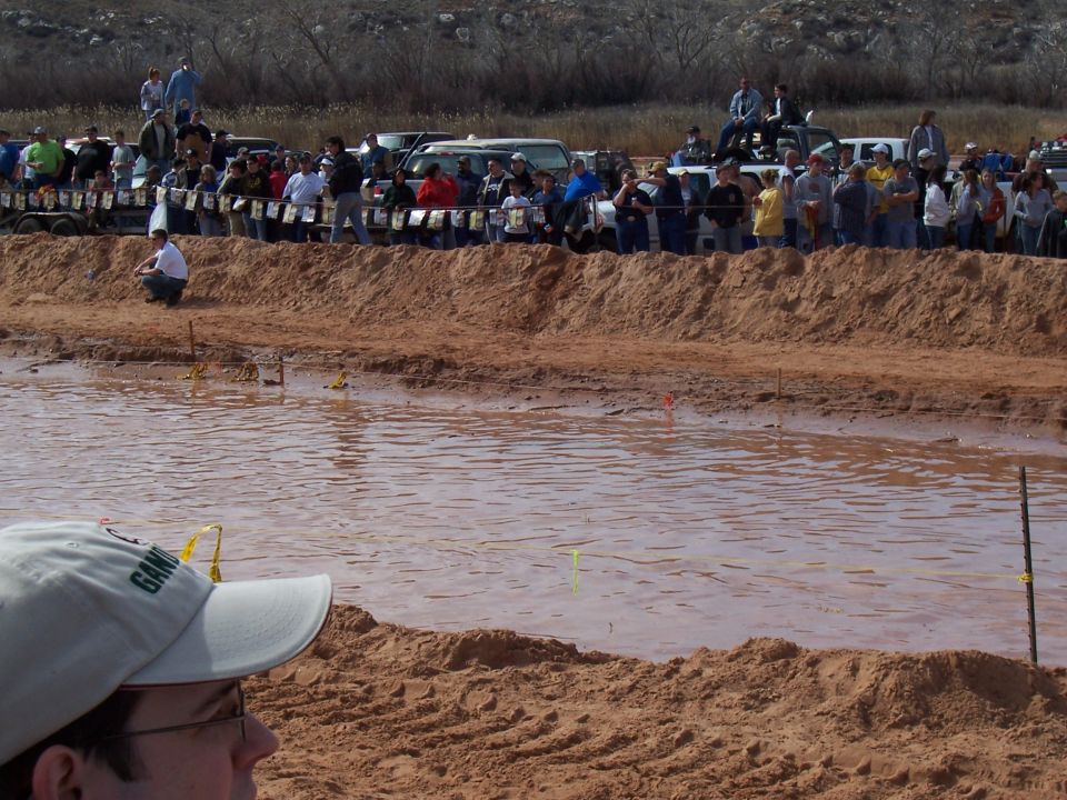 Spectators watch a mud bog event from a sandy embankment overlooking a muddy pool. Several vehicles are parked in the background