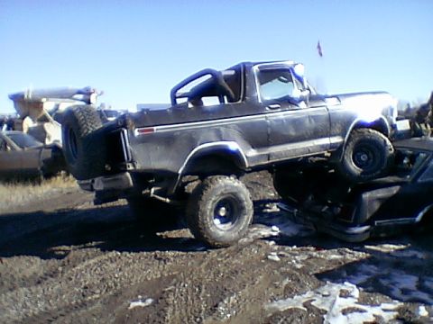 1979 Ford Bronco - This lifted Ford Bronco is parked on top of another vehicle in a junkyard setting.
