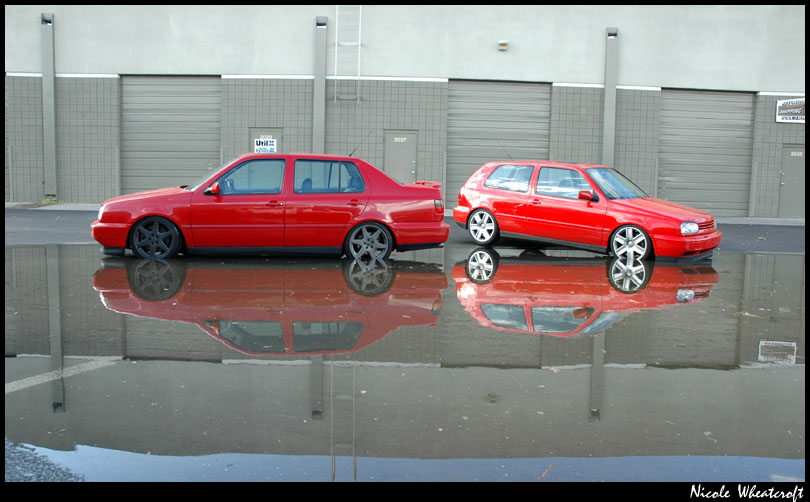 Two red Volkswagen Jettas, one sedan and one hatchback, parked side-by-side with their reflections in a wet lot