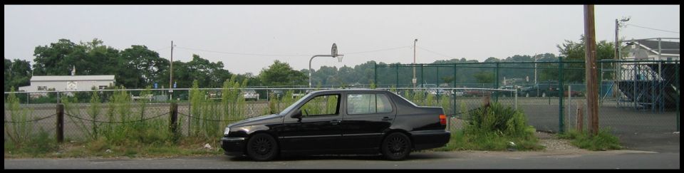 Black sedan parked on a dirt lot next to a chain-link fence. The vehicle has tinted windows and black wheels