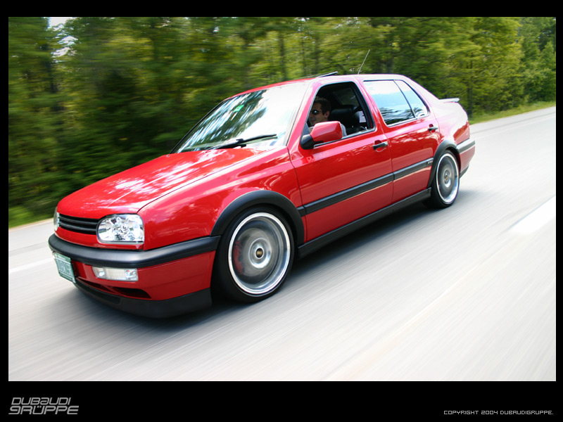 Red sedan driving on a road, front 3/4 view, motion blur on wheels