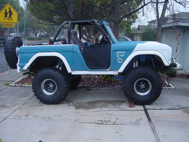 Side view of lifted teal early Ford Bronco with white fenders, no doors, roll cage, and big off-road tires in a driveway