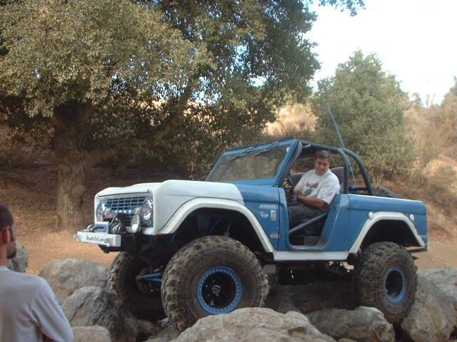 Lifted blue-and-white doorless Ford Bronco with big tires and roll cage climbing rocks on a trail