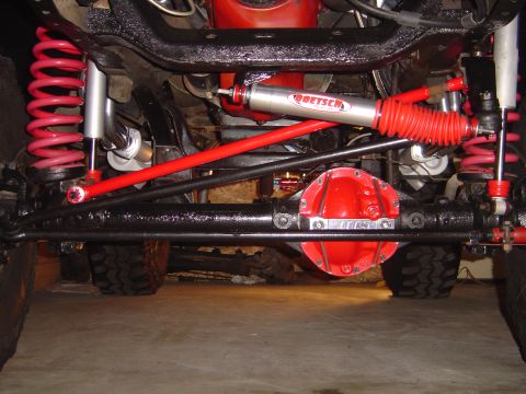 1978 Ford Bronco - Front underbody view of a lifted 4x4 solid front axle with a red aftermarket differential cover, black tie rod and drag link, red-painted r…