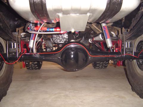 1978 Ford Bronco - Rear underbody view of a lifted truck showing a gloss-black solid rear axle housing with a red-painted brake line running across it, leaf s…