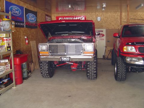 1978 Ford Bronco - Front view of a lifted red square-body Ford Bronco in an OSB-lined shop with its hood raised, showing the chrome grille and bumper with a T…