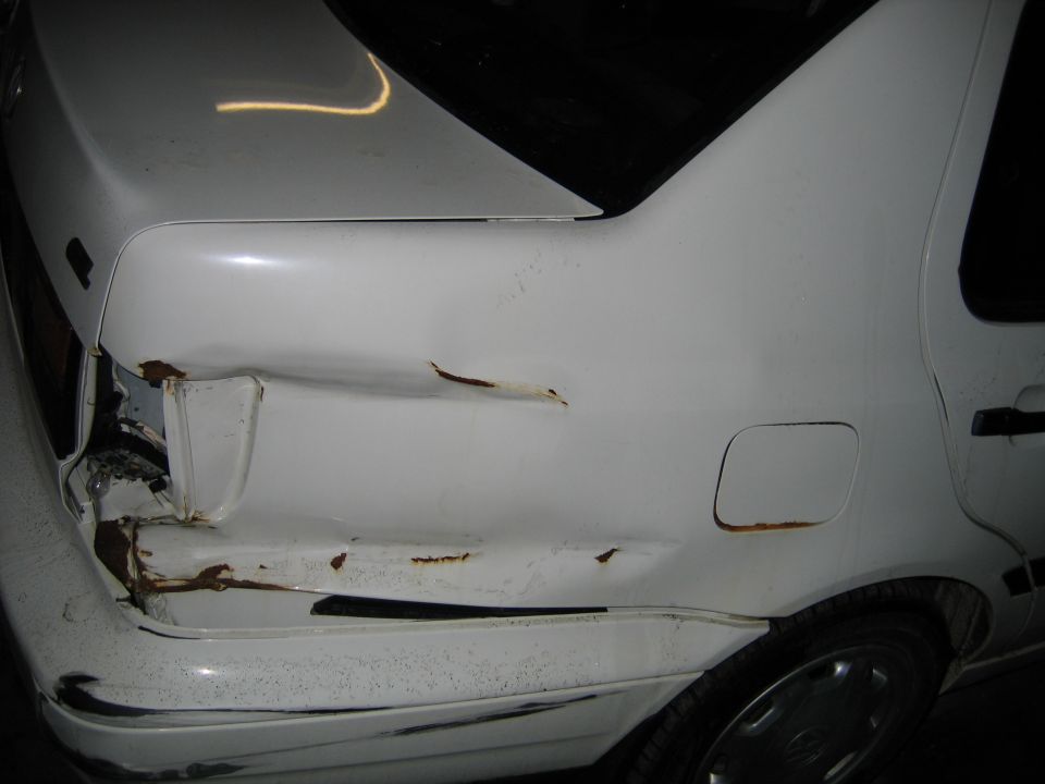 White sedan's rear passenger side has significant damage and rust near the taillight and along the body panel