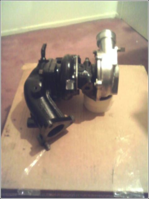 1989 Mazda MX-6 - A turbocharger unit, a black exhaust manifold and a silver compressor housing, is displayed on a cardboard surface.