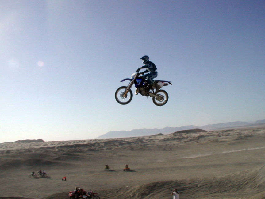 Rider jumping a blue dirt bike high over desert dunes with mountains in the distance