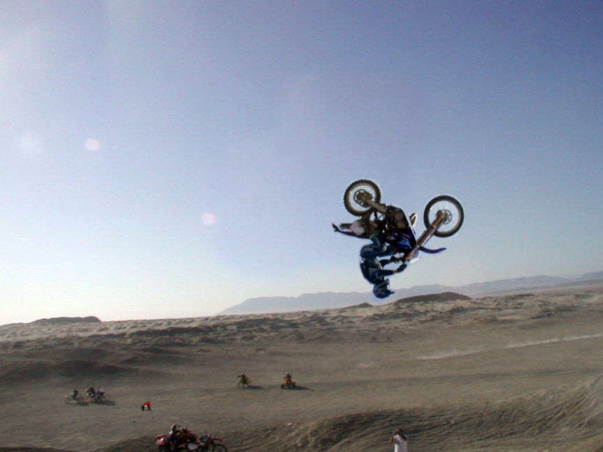 Motocross rider mid-air on a dirt bike over desert dunes under blue sky