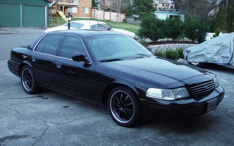 1998 Ford Crown Victoria - A black sedan is parked on a concrete driveway with a covered vehicle in the background.