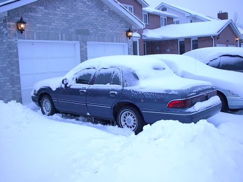 1985 Mercury Grand Marquis - A dark blue sedan, likely a 1996 Mercury Grand Marquis, sits buried in snow next to a garage.