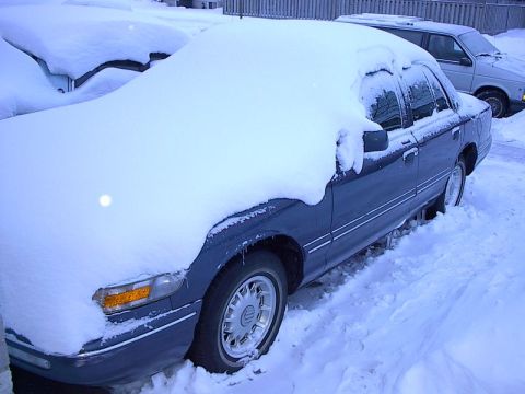 1985 Mercury Grand Marquis - A dark blue sedan is heavily covered in snow, with another vehicle visible in the background.