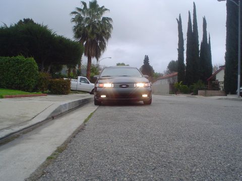 1993 Ford Taurus - A dark 1993 Ford Taurus is parked on a residential street, facing the camera with its headlights illuminated.