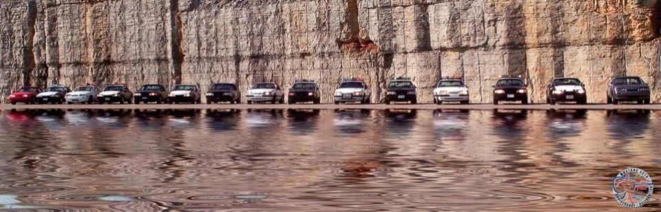 Lineup of Fox-body Mustang police cars parked along a rock cliff, reflected in water