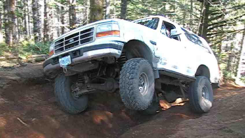 White lifted Ford Bronco SUV flexing over dirt obstacle on wooded off-road trail