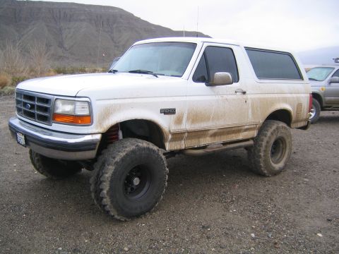 1993 Ford Bronco - A white 1993 Ford Bronco, modified for off-road use, is seen parked on a dirt lot with another vehicle in the background.