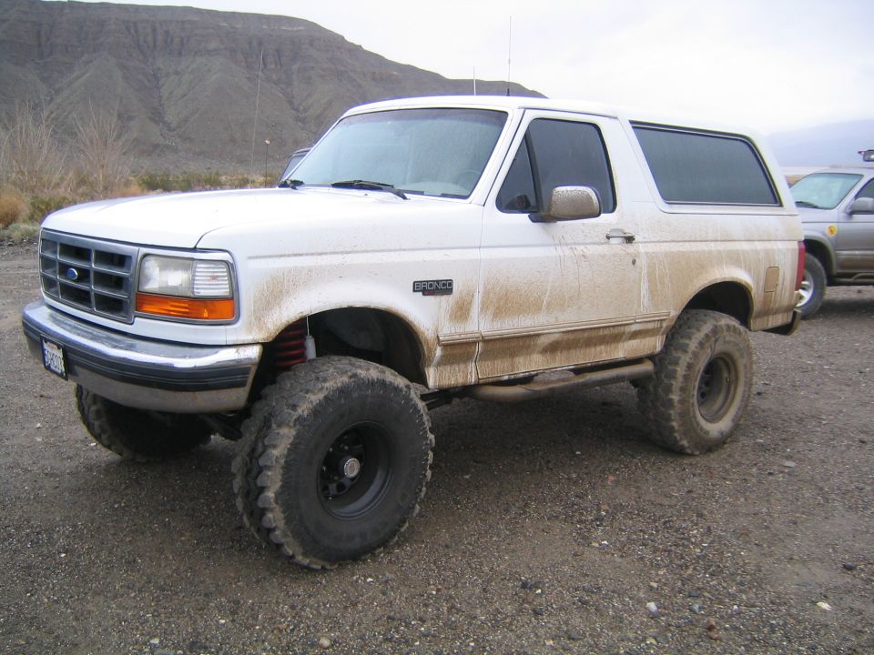 Lifted white Ford Bronco covered in mud spray on big tires in a desert gravel lot