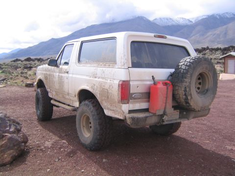 1993 Ford Bronco - A white 1993 Ford Bronco equipped with a spare tire and two red gas cans is parked in a dusty, mountainous landscape.