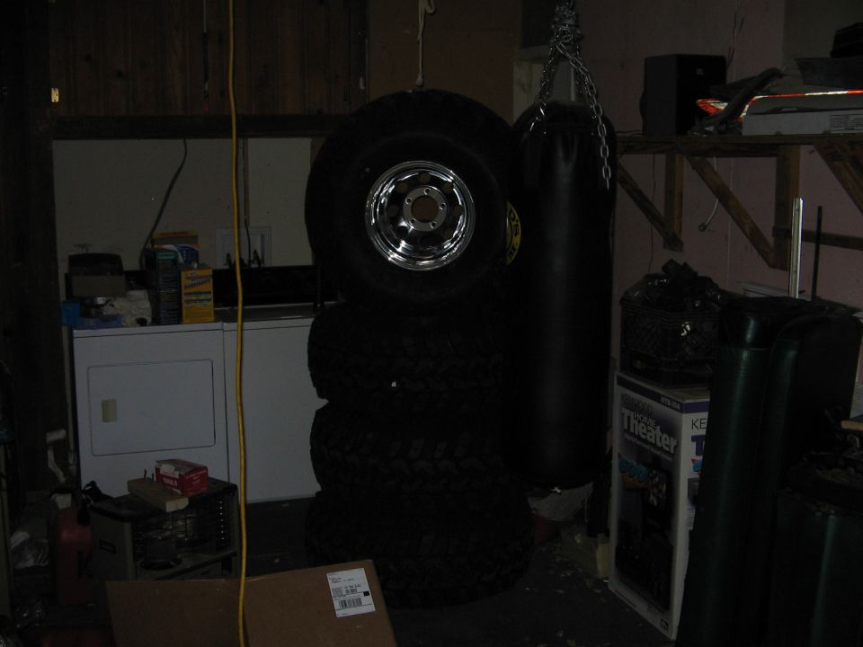 Stack of four tires, one with a chrome rim, next to a black punching bag in a cluttered garage