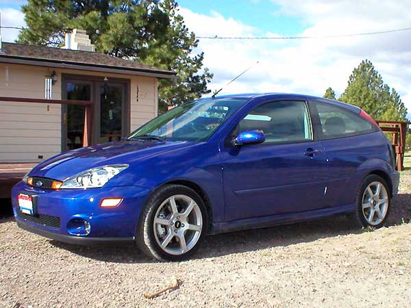 Blue Ford Focus 3-door hatchback with silver alloy wheels parked on gravel by a house and pine trees