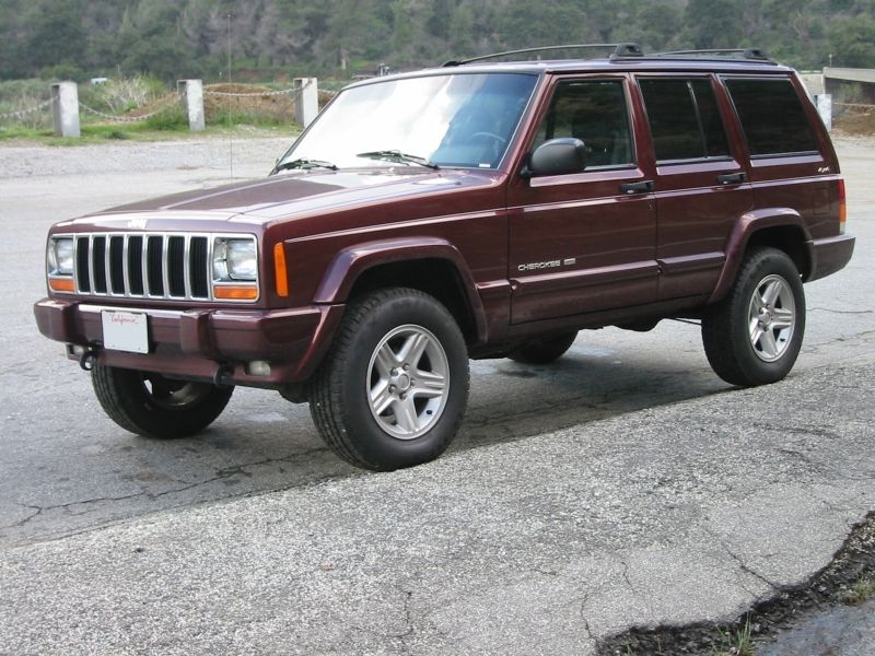 Dark maroon Jeep Cherokee XJ 4-door SUV, front three-quarter view, parked on cracked pavement with trees behind.