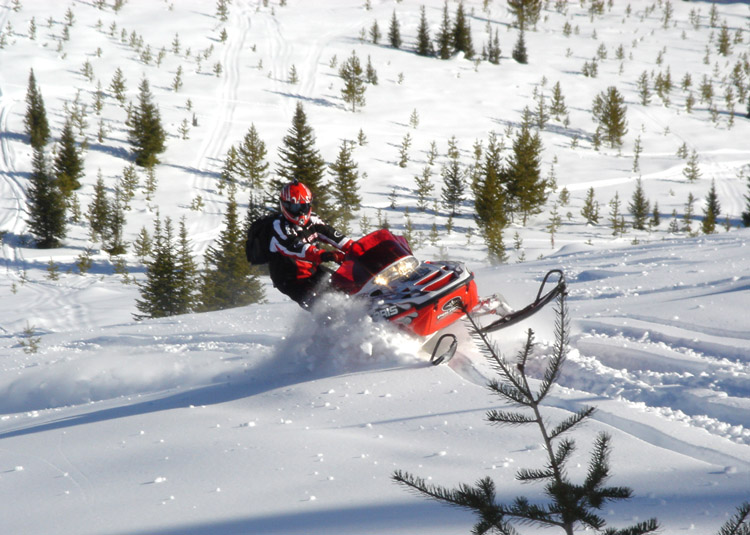 Rider on red snowmobile carving through deep snow in a forested mountain field