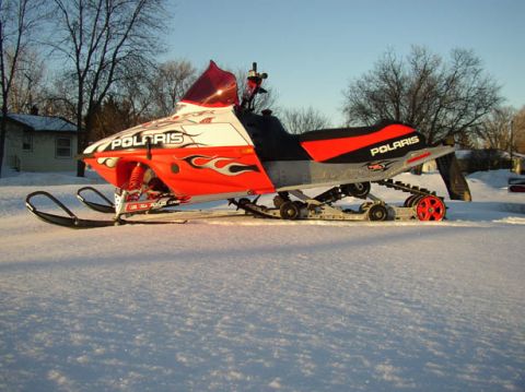 2003 Polaris 600 PRO X - A 2003 Polaris snowmobile sits in the snow, displaying its side profile with vibrant orange and white graphics.