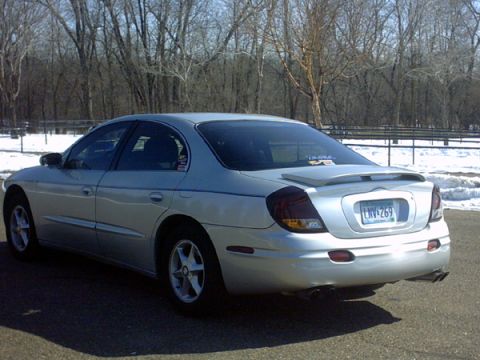 2002 Oldsmobile Aurora - A silver sedan is parked outdoors with trees and snow in the background. The vehicle has a spoiler and dual exhaust tips.