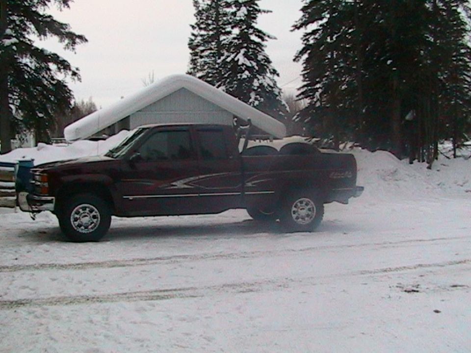 Maroon pickup truck parked in a snowy area with a house and trees in the background