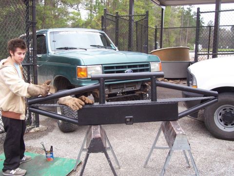 1989 Ford Bronco - A man works on a custom front bumper for a green 1989 Ford Bronco, with another vehicle visible to the right.