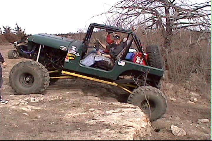 Green off-road Jeep climbing over rocks with a person in the driver's seat. Another vehicle is visible in the background