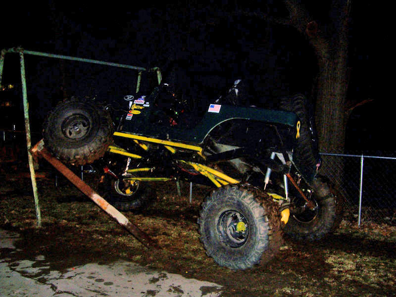 Modified Jeep with large tires stuck on a metal bar, next to a tree and fence