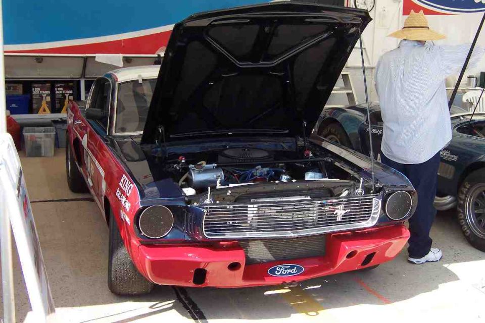 Front view of a 1965 Ford Mustang race car with hood open, red and blue paint, and racing decals. Another car is visible to the right