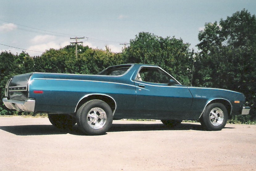Side view of a blue Ford Ranchero pickup with chrome five-spoke wheels parked on gravel by trees