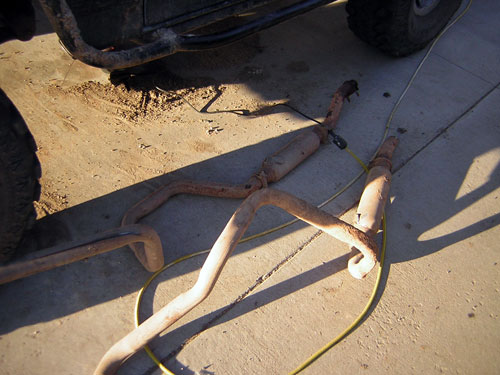 Exhaust pipes and mufflers lying on concrete next to a vehicle's tire and frame
