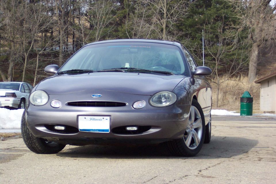 Gray Ford Taurus from the front-right, with a white car in the background