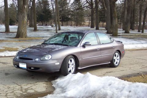 1996 Ford Taurus - A gray sedan is parked on a road with snow and ice on the ground, surrounded by trees.